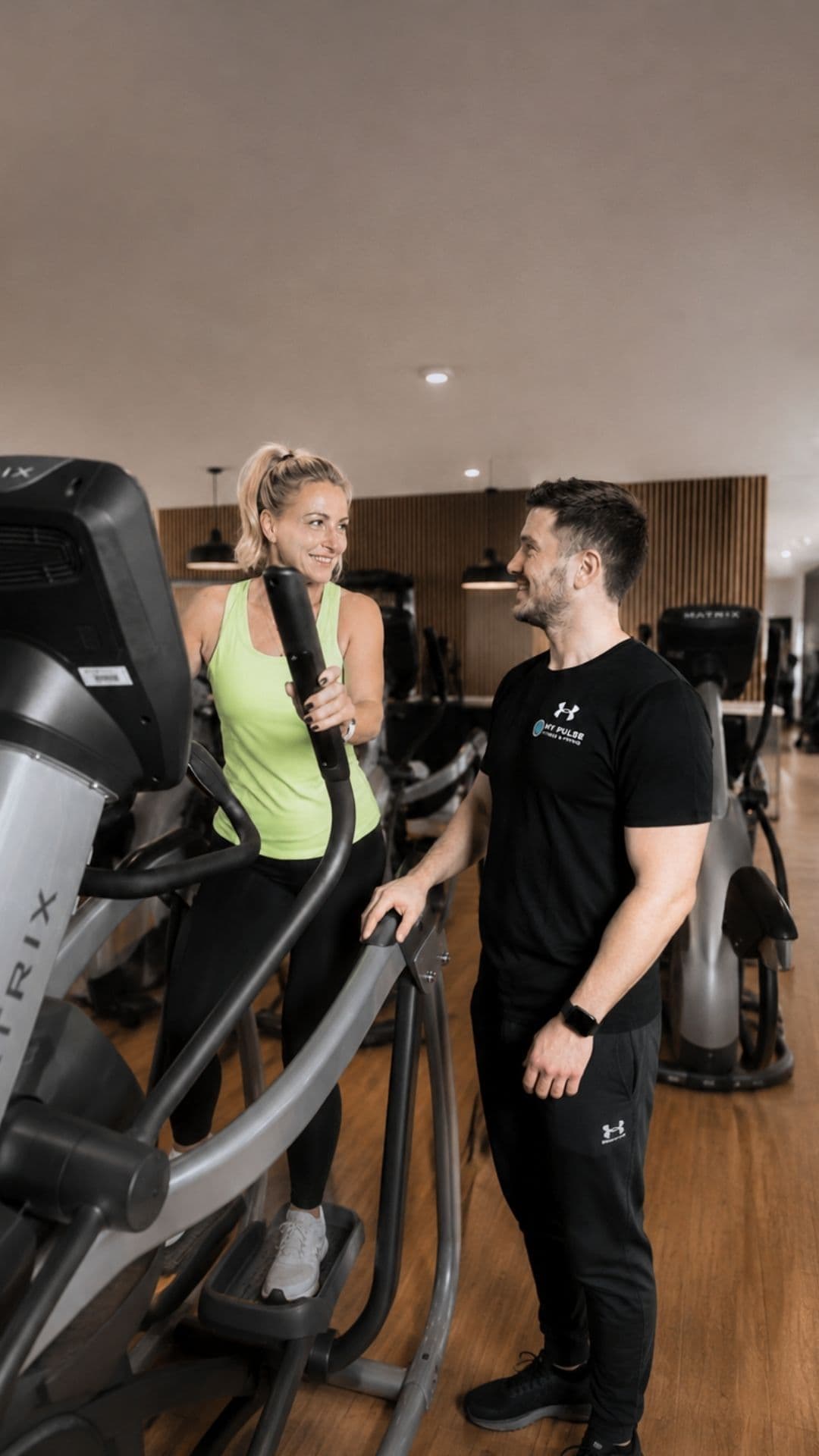 Mitglied trainiert am Crosstrainer, ein Trainer steht daneben im Gespräch