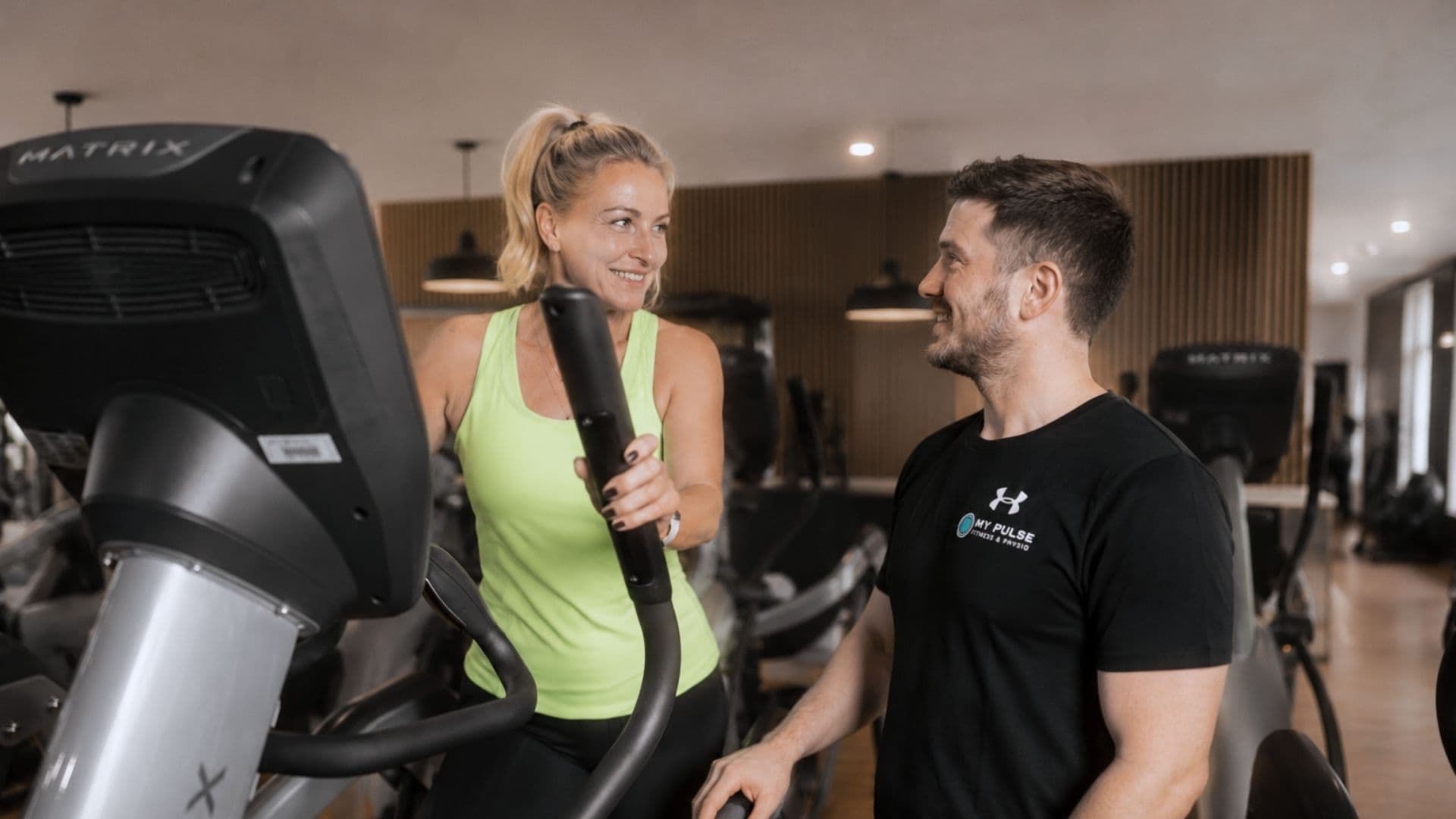 Mitglied trainiert am Crosstrainer, ein Trainer steht daneben im Gespräch